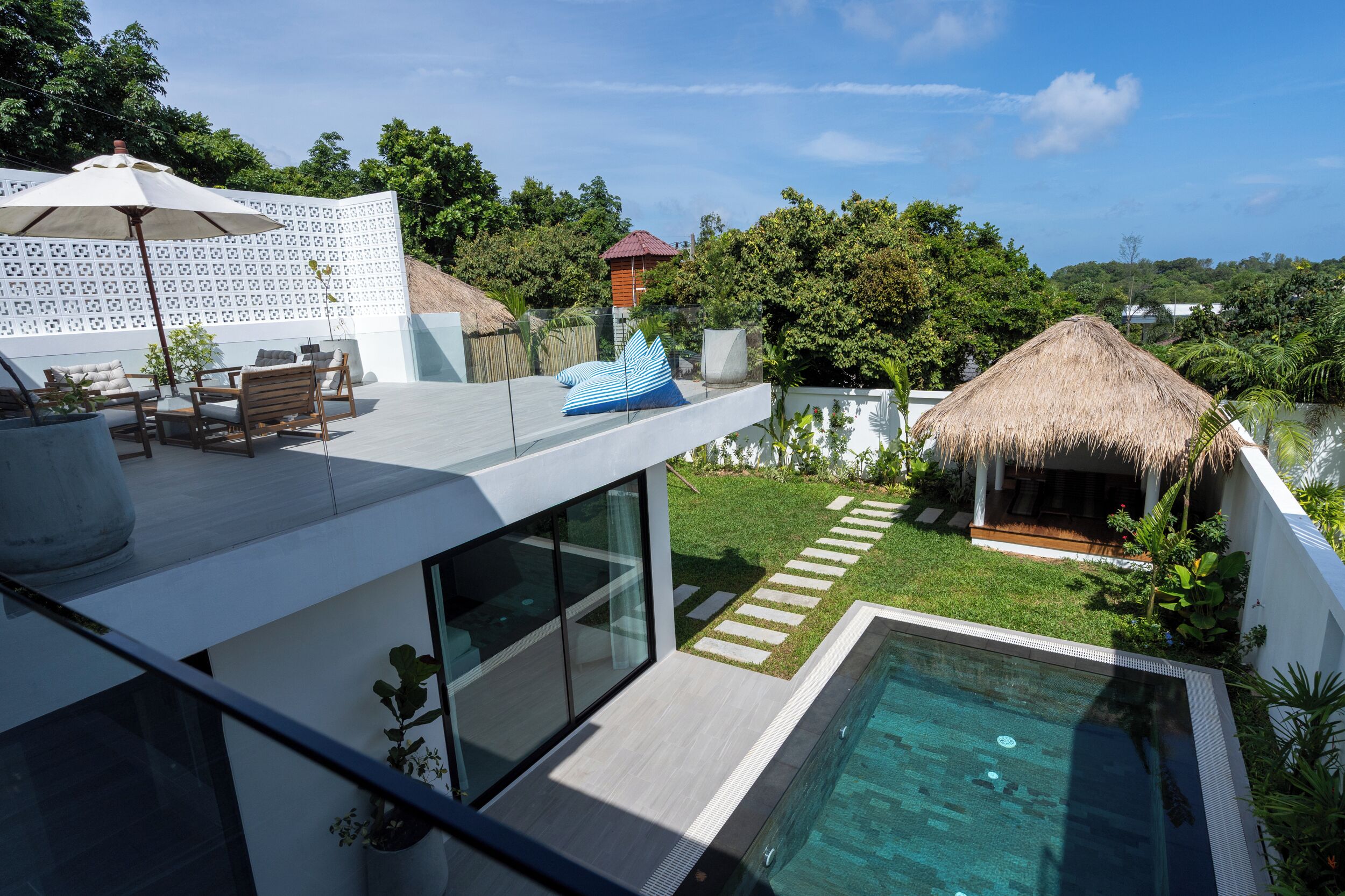 A modern outdoor pool area with a glass balustrade, a thatched-roof structure, and a seating area with a table and chairs.