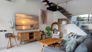 A modern living room with a large flat-screen TV, a wooden coffee table, a grey sofa, and a staircase with a glass railing. - The Guu Villas (Ko Lanta)