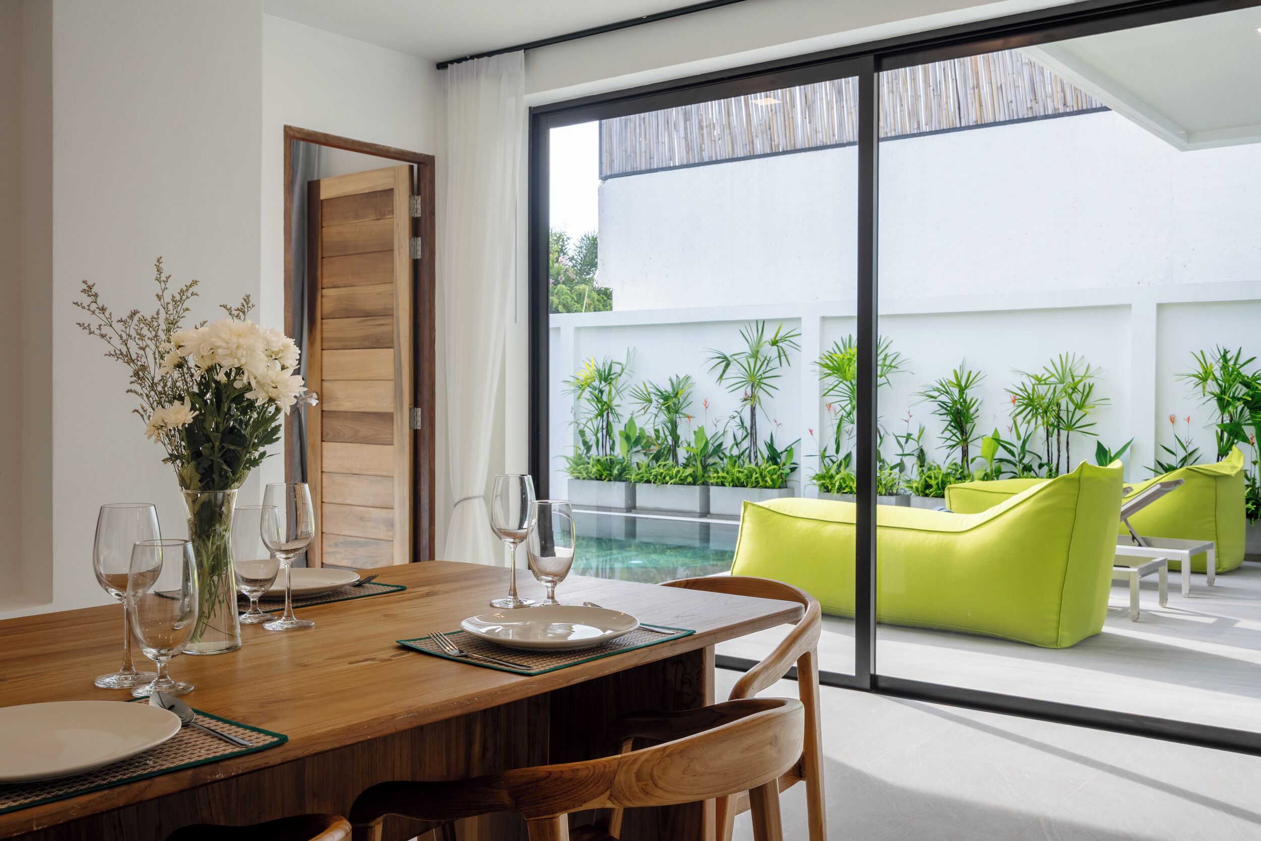 A dining area with a wooden table, chairs, and a vase of flowers. There is a sliding glass door leading to an outdoor patio with green plants and a yellow sofa.