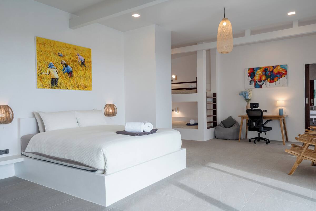 A modern bedroom with a large bed, a desk, and a chair. There are paintings on the walls and a hanging light fixture.