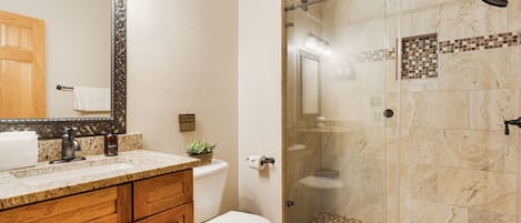 Un baño con tocador de madera, inodoro, ducha con mampara de vidrio y suelo de baldosas.