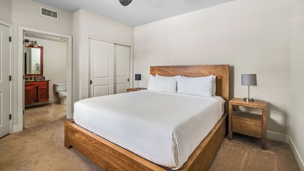 Un dormitorio con cama de madera, ropa de cama blanca, una mesita de noche con lámpara y un baño visible a través de una puerta abierta.