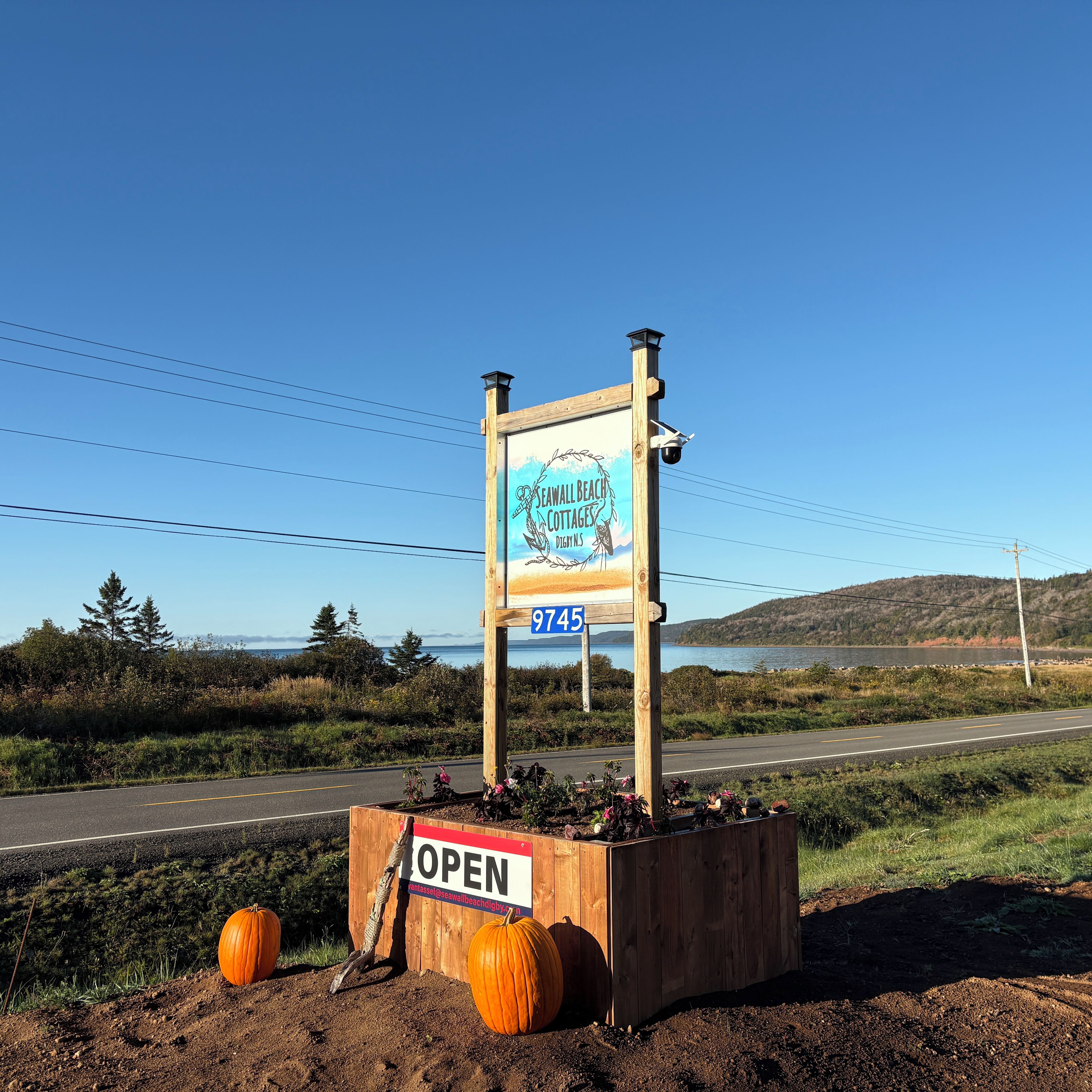 Un panneau en bois, orné du logo d’un complexe de chalets de plage et d’une adresse électronique de contact, est placé au bord de la route, accompagné de deux citrouilles et d’un jardinière en bois.