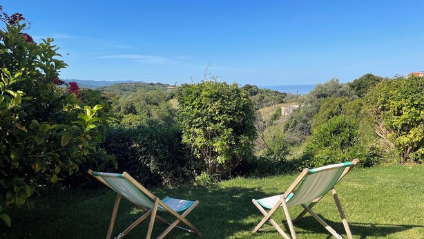 Two wooden folding chairs on a grassy lawn with a view of a hilly landscape and the sea in the distance.
