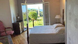 A bedroom with a large bed, wooden floor, a desk with a chair, and a view of a green landscape through an open door.