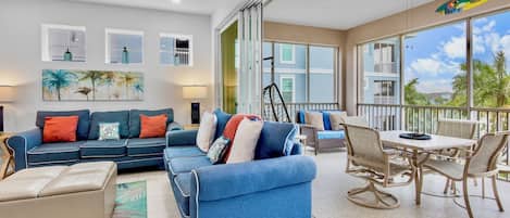 A spacious living area with a blue sofa, a dining table with chairs, and a balcony with a view of palm trees.