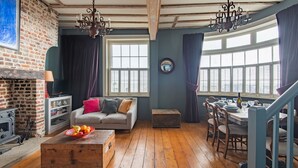 Ein rustikales Wohnzimmer mit sichtbaren Holzbalken, einer Ziegelwand, einem Kamin und einem Essbereich mit großem Fenster.