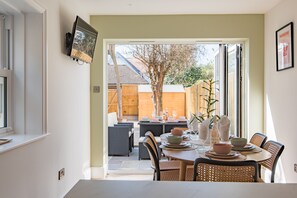 A dining area with wicker furniture, a table set with plates, bowls, and glasses, and a view of a backyard with a wooden fence and trees. - Brook Cottage, Whitstable (Whitstable)