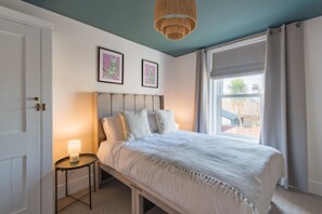 A bedroom with a bed, a bedside table with a lamp, two framed pictures on the wall, and a window with curtains. - Brook Cottage, Whitstable (Whitstable)