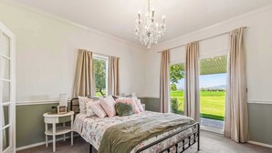 A bedroom with a bed, a bedside table, a chandelier, and a view of a green landscape through large windows.