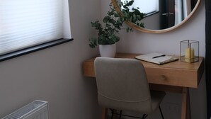 Une pièce d’angle avec un bureau en bois, un miroir rond, une plante en pot, une chaise, un radiateur et une fenêtre avec des stores.