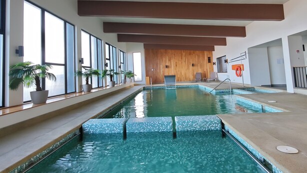 Une piscine intérieure à l’eau d’un bleu cristallin, avec des poutres en bois au plafond et des plantes en pot disposées le long des fenêtres.