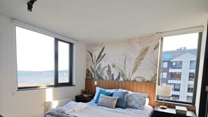 A bedroom with a bed, a nightstand, a lamp, a window with a view of the sea, and a ceiling light. - Apartment in Puerto Natales, cozy and with all the comforts you need. (Puerto Natales)