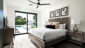Une chambre moderne avec un grand lit, un ventilateur de plafond, une table de chevet avec une lampe, et une vue sur une piscine par le biais d’une porte vitrée.