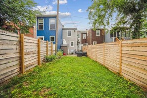 A narrow backyard with a wooden fence, a grassy area, and a row of houses in the background.