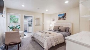 A bedroom with a bed, bedside table, chair, and a view of the kitchen.