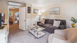 A living room with a sofa, a coffee table, and a TV. There is a door leading to another room, and a painting on the wall.