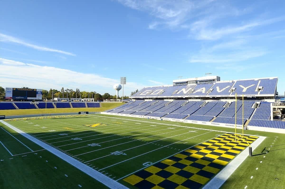 A football stadium with "50 NAVY" branding, a large scoreboard, and empty blue seating.