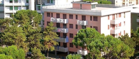 A hotel with a pool surrounded by trees, a view of the sea, and multiple balconies with air conditioning units.