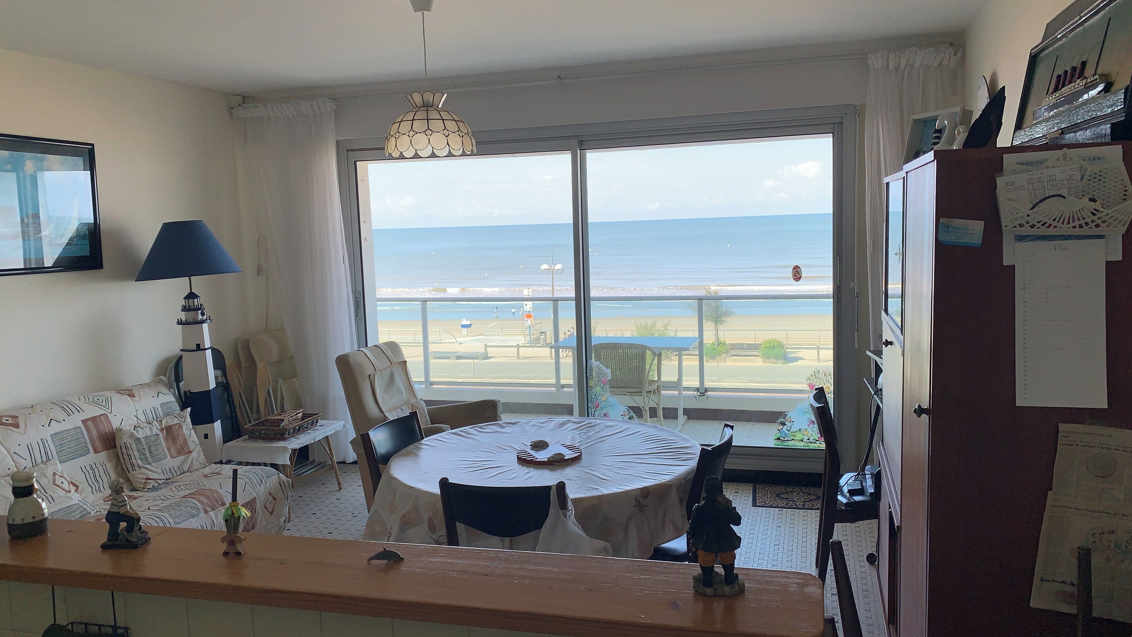 Une pièce dotée d’une grande fenêtre donnant sur la plage et l’océan. On y trouve une table ronde avec des chaises, une lampe et une petite étagère ornée d’objets décoratifs.