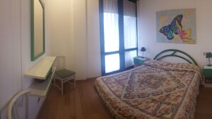 A bedroom with a bed, a desk, a chair, a window with curtains, a nightstand, and a wall painting. - Bilo in residence+beach from 01/06/25 to 30/09/25 (Capanne-Prato-Cinquale)