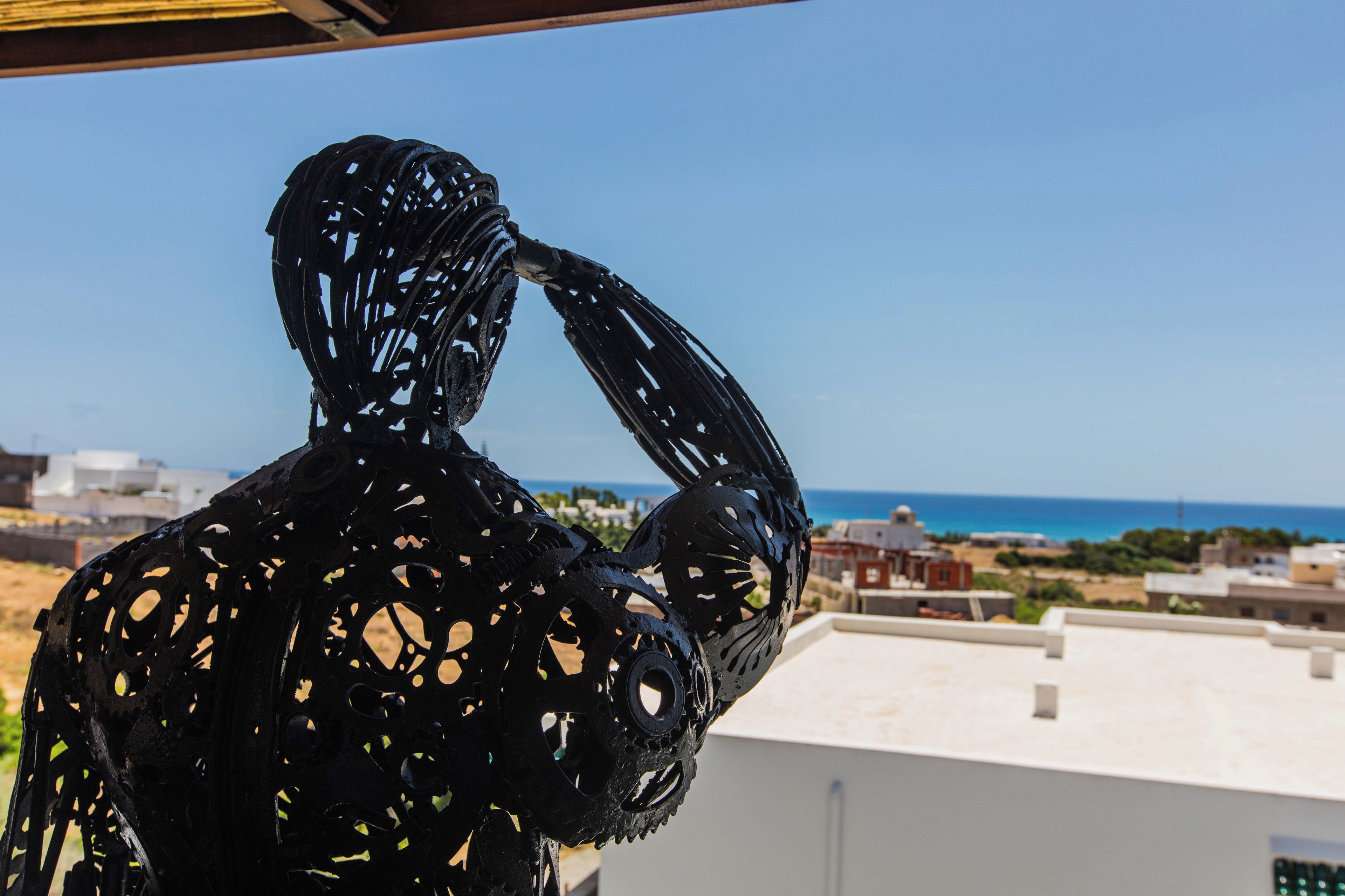 A sculpture of a human figure with a complex, abstract design, set against a backdrop of a clear sky and a distant coastal landscape.
