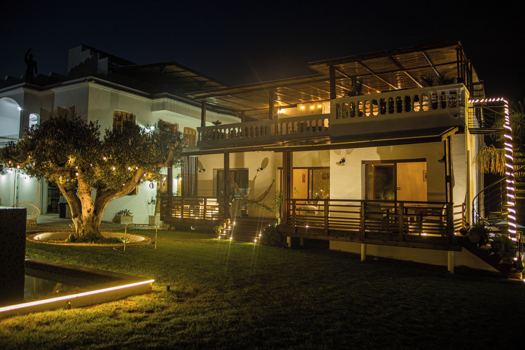 Une maison à deux étages avec un balcon, un arbre dans la cour et des guirlandes lumineuses.