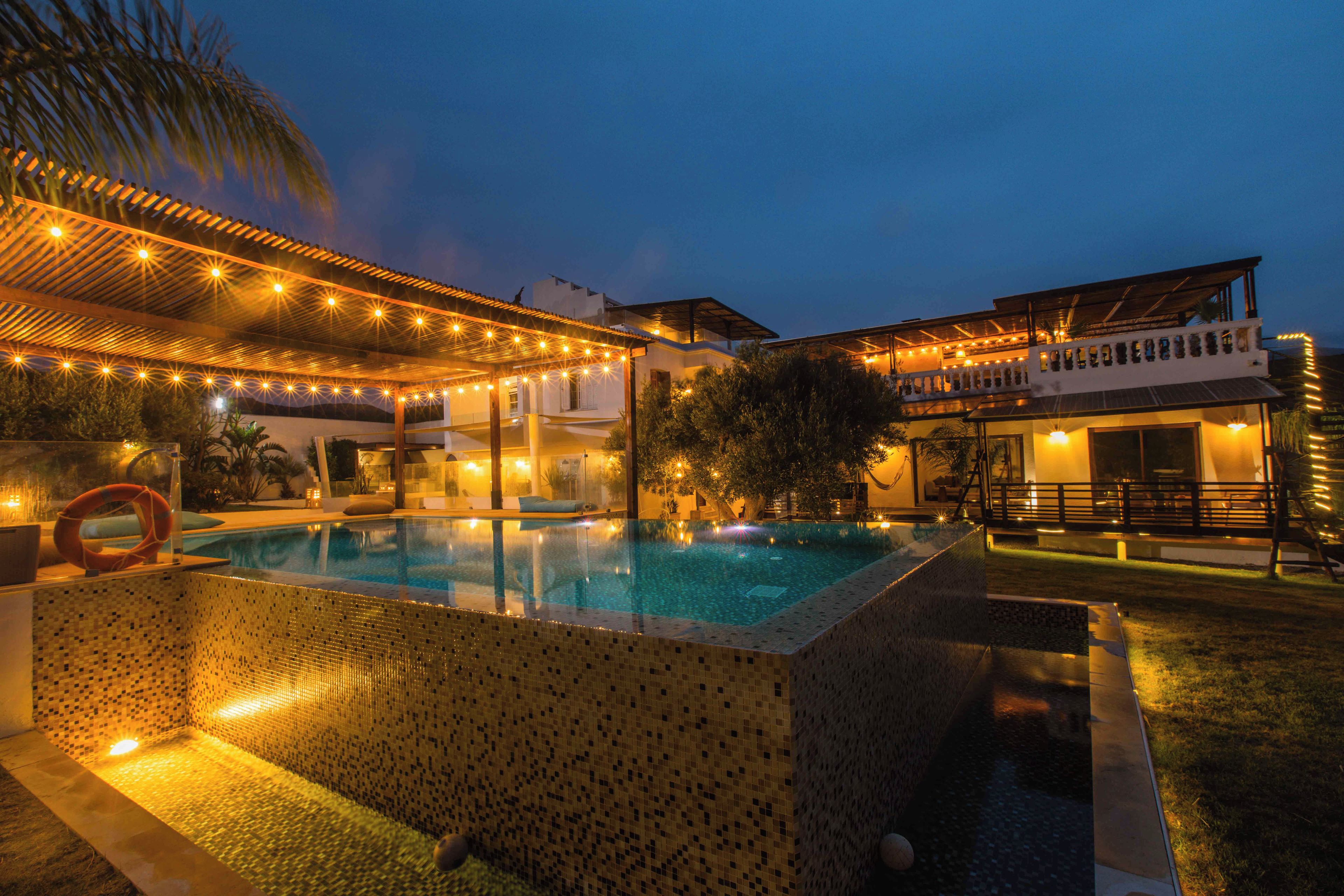 A swimming pool with a tiled edge, surrounded by a covered patio area with string lights.