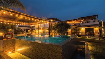 Une piscine dont le bord est revêtu de carreaux, entourée d’un patio abrité et illuminé de guirlandes lumineuses.