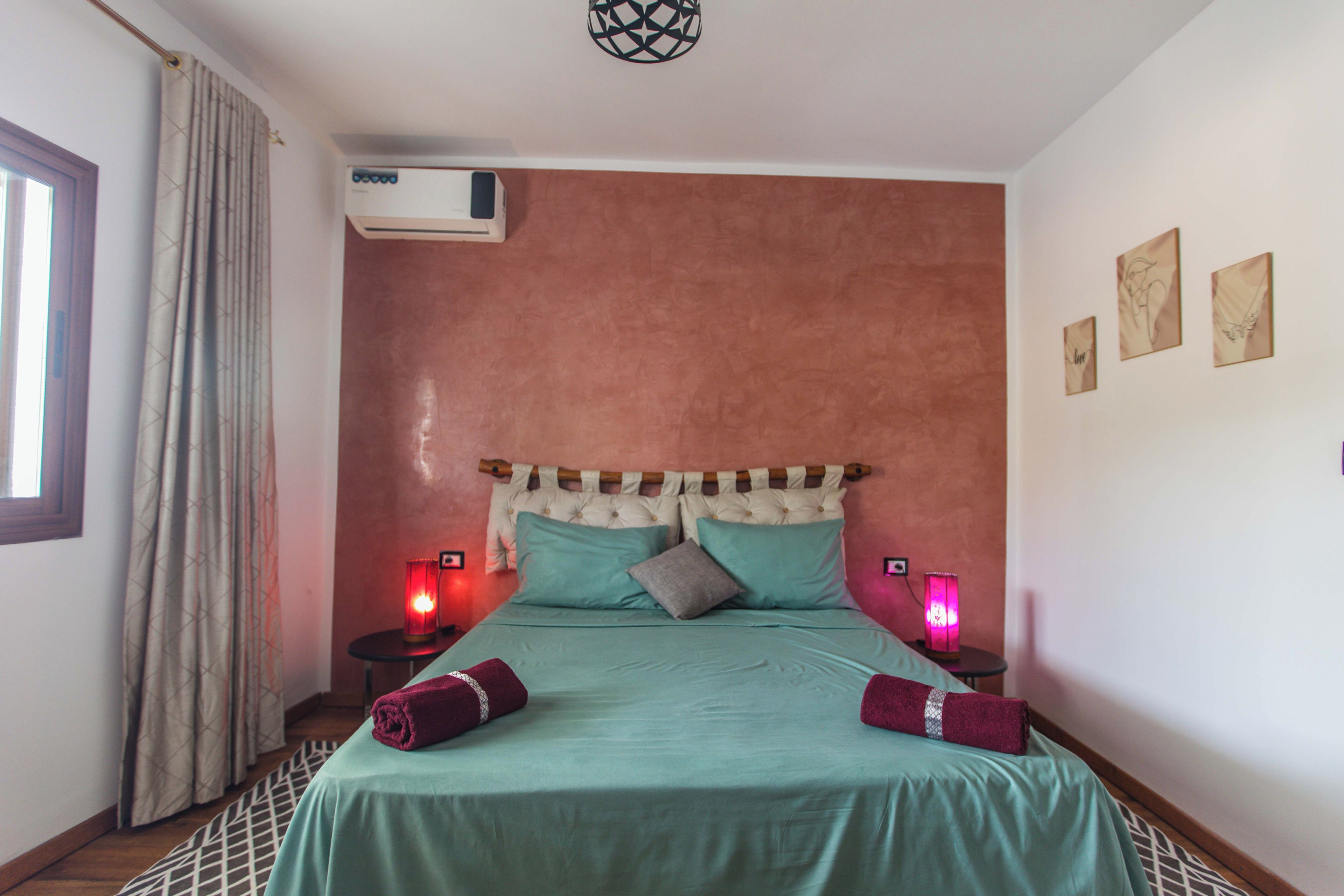 A bedroom with a bed, two bedside lamps, a window with curtains, and an air conditioning unit.