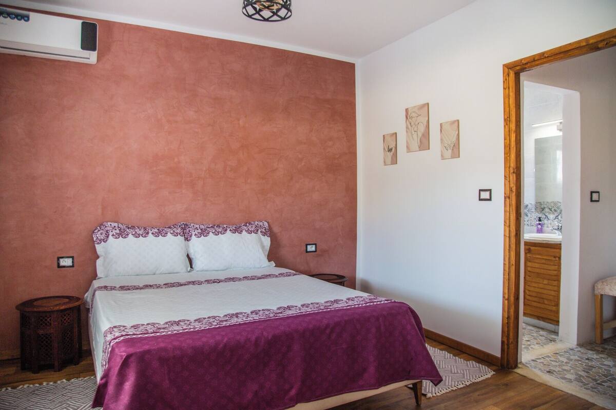 A bedroom with a bed, a wooden floor, a small round table, and a door leading to a bathroom.