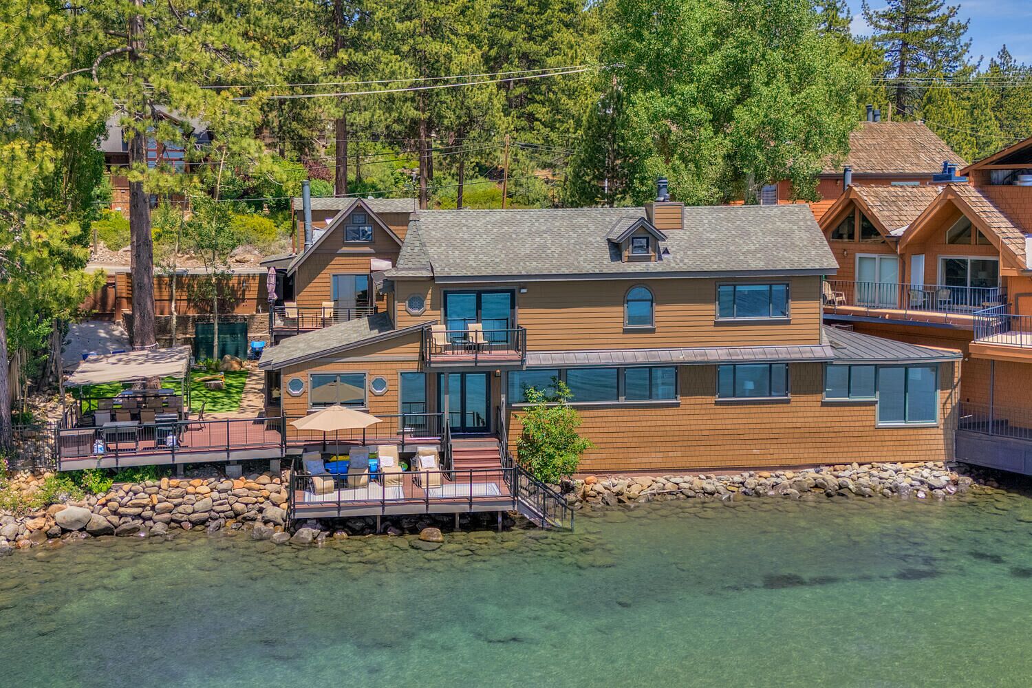 Une maison à plusieurs étages dotée d’une terrasse surplombant un plan d’eau aux eaux claires, le tout entouré d’arbres.