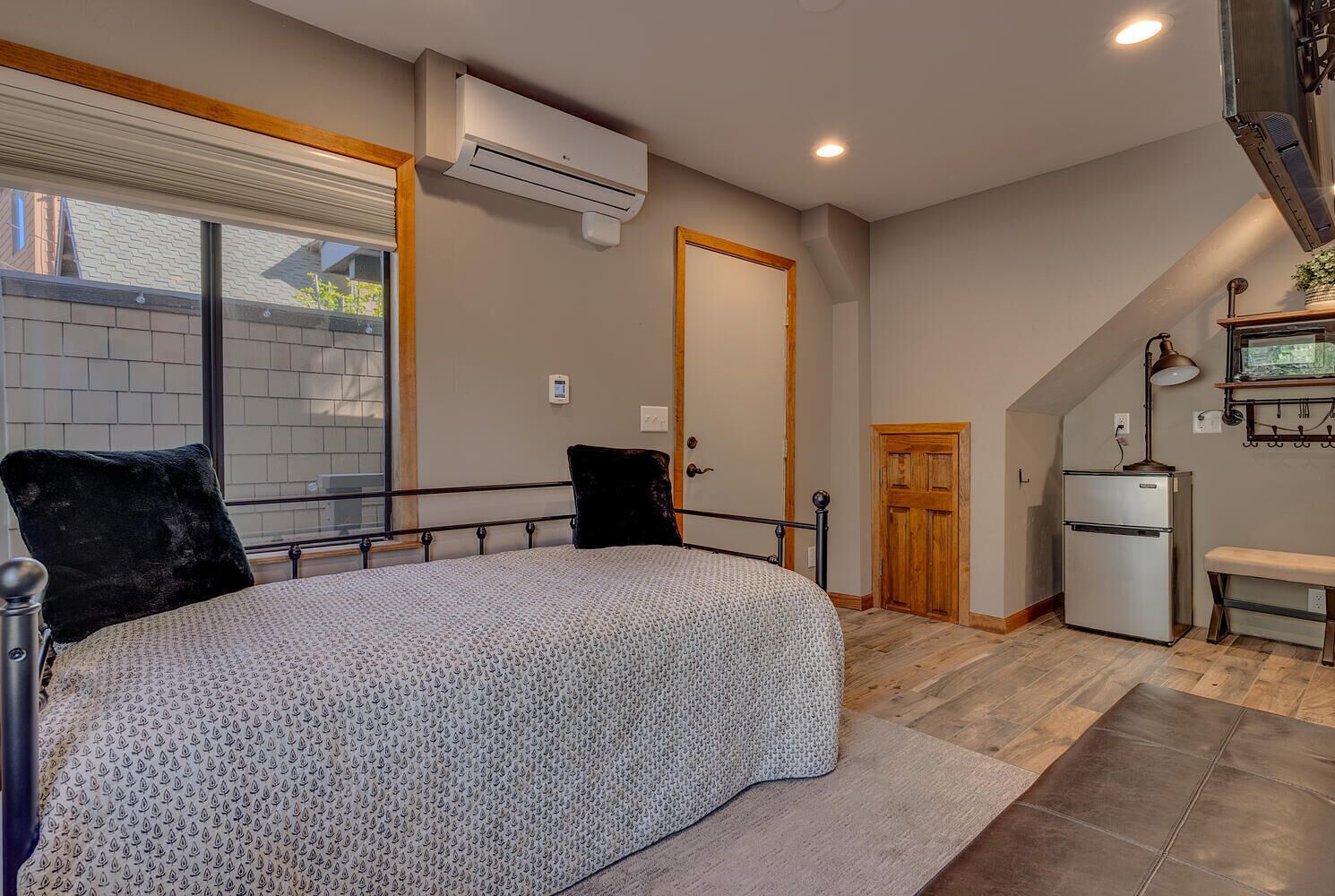 A bedroom with a bed, a window, an air conditioner, a refrigerator, and a bench.