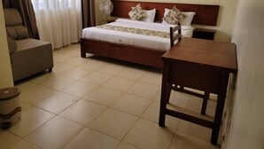 Una habitación de hotel con una cama, una mesita de noche de madera, una silla, una pequeña mesa y un ventilador de pie.