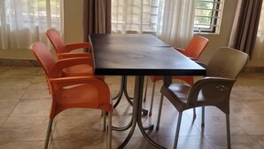 Un comedor con una mesa rectangular rodeada por seis sillas, algunas naranjas y otras grises, en una habitación con suelo de baldosas y ventanas grandes con cortinas.
