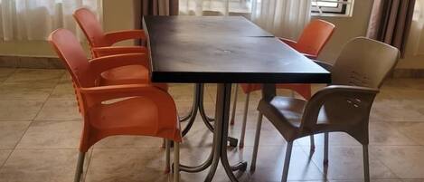 Un comedor con una mesa rectangular rodeada por seis sillas, algunas naranjas y otras grises, en una habitación con suelo de baldosas y ventanas grandes con cortinas.