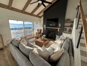A spacious living room with a large gray sofa, a fireplace, and a view of the sea through large windows. - The Landing: 3BR/3BA Waterfront Condo in Charming Boyne City. (Boyne City)