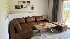 En moderne stue med en brun lædersofa, et træbord og et stort vindue med gardiner.