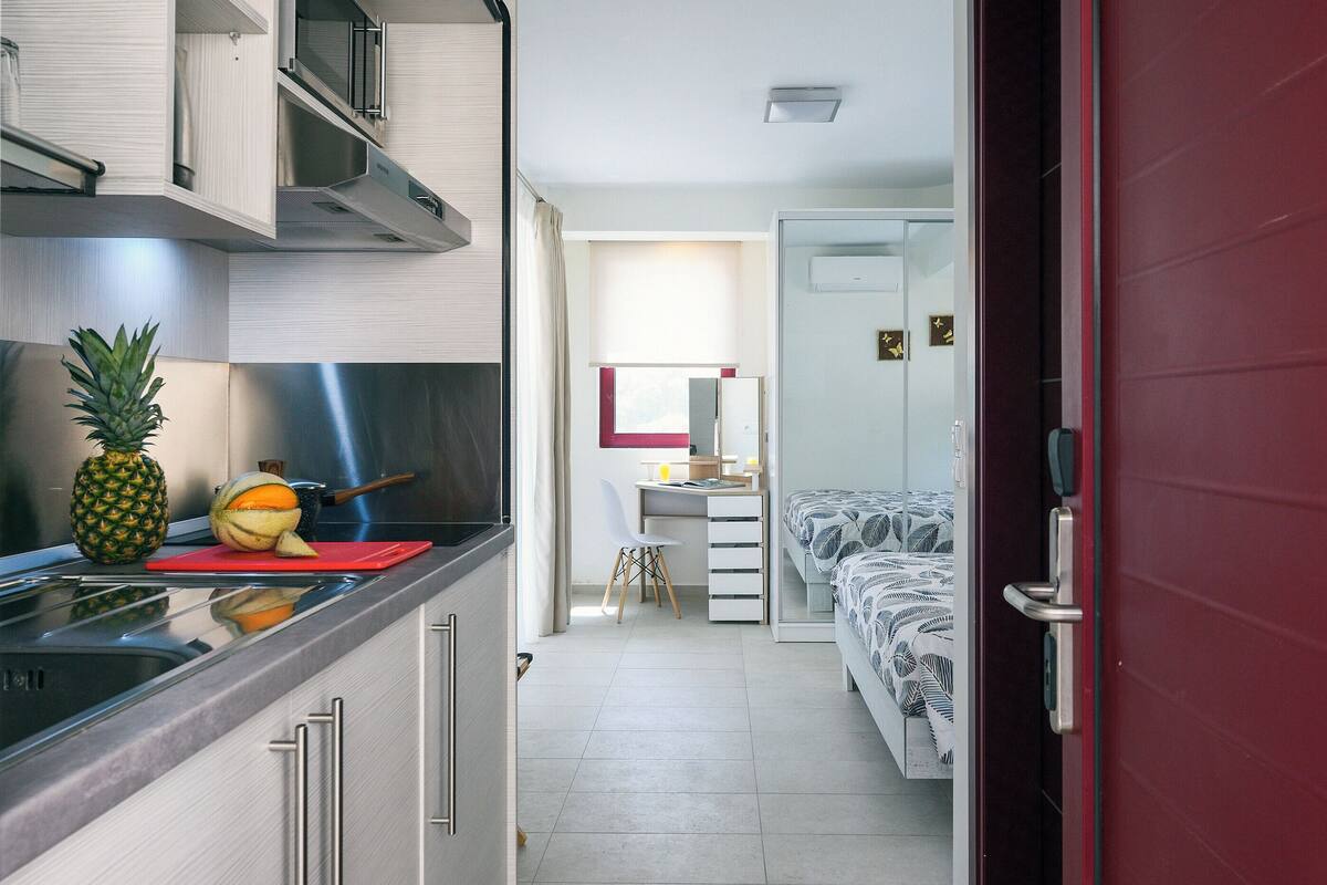 Eine kompakte Küche mit modernen Geräten, ein Essbereich mit einem roten Tisch und ein Schlafzimmer mit Bett und Nachttisch.