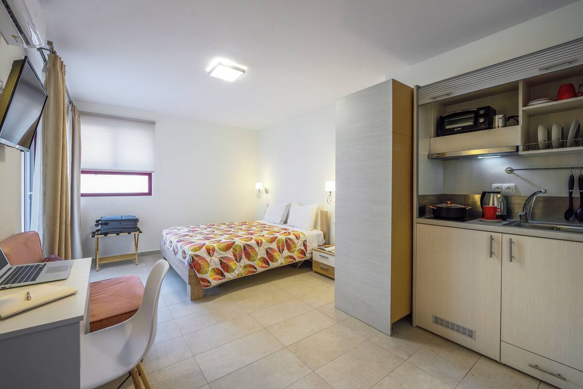 Ein kompaktes Hotelzimmer mit Bett, Schreibtisch, einem Laptop, einem kleinen Tisch und einer Küchenzeile.