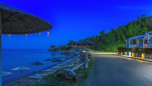 Eine Küstenstraße mit Strandstühlen und Sonnenschirmen, einem Gebäude mit Reetdach und Häusern mit Balkonen, die auf das Meer blicken.