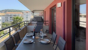 Un balcon avec une table dressée pour deux personnes, une bouteille de vin et un verre. Le balcon est doté d’un mur rouge et offre une vue sur des bâtiments et des arbres en arrière-plan.