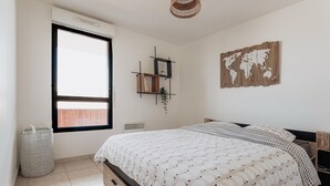 Une chambre avec un grand lit, une fenêtre, une carte du monde accrochée au mur et une petite étagère avec des livres et une plante.