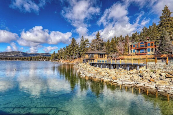 A lakeside property with a wooden dock, a stone pier, and a large house with multiple windows and a green roof.