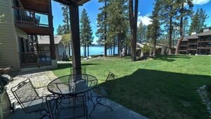 A covered patio with a round table and chairs, a grassy lawn, and a view of a lake and tall trees. - Lakefront Condo in Kings Beach-#37 (Kings Beach)