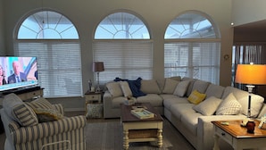 A living room with a sofa, armchair, and coffee table, large arched windows, and a television.