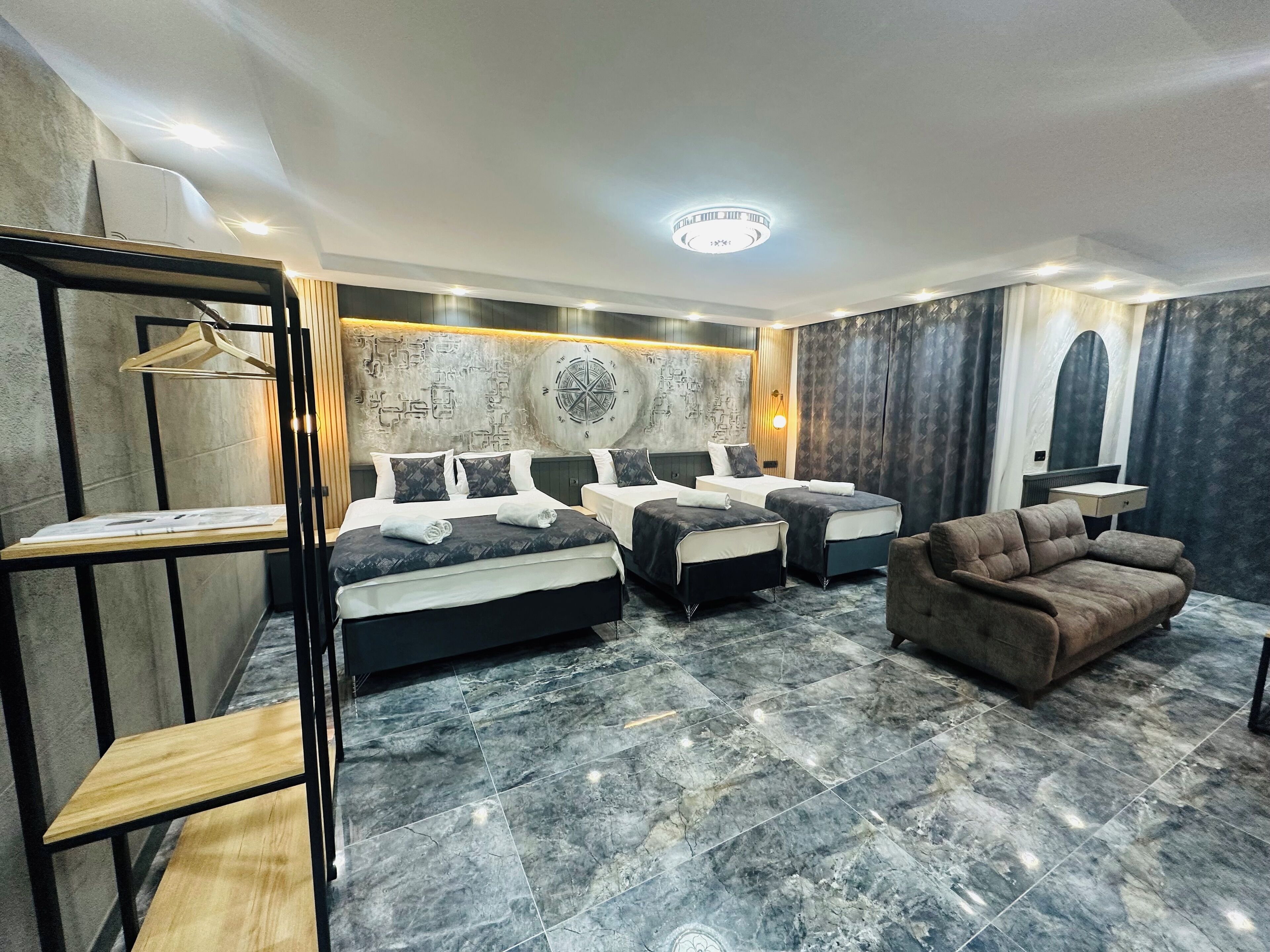 Moderni hotellihuone, jossa on kolme sänkyä, sohva, puinen hylly ja marmorilaatoilla päällystetty lattia.