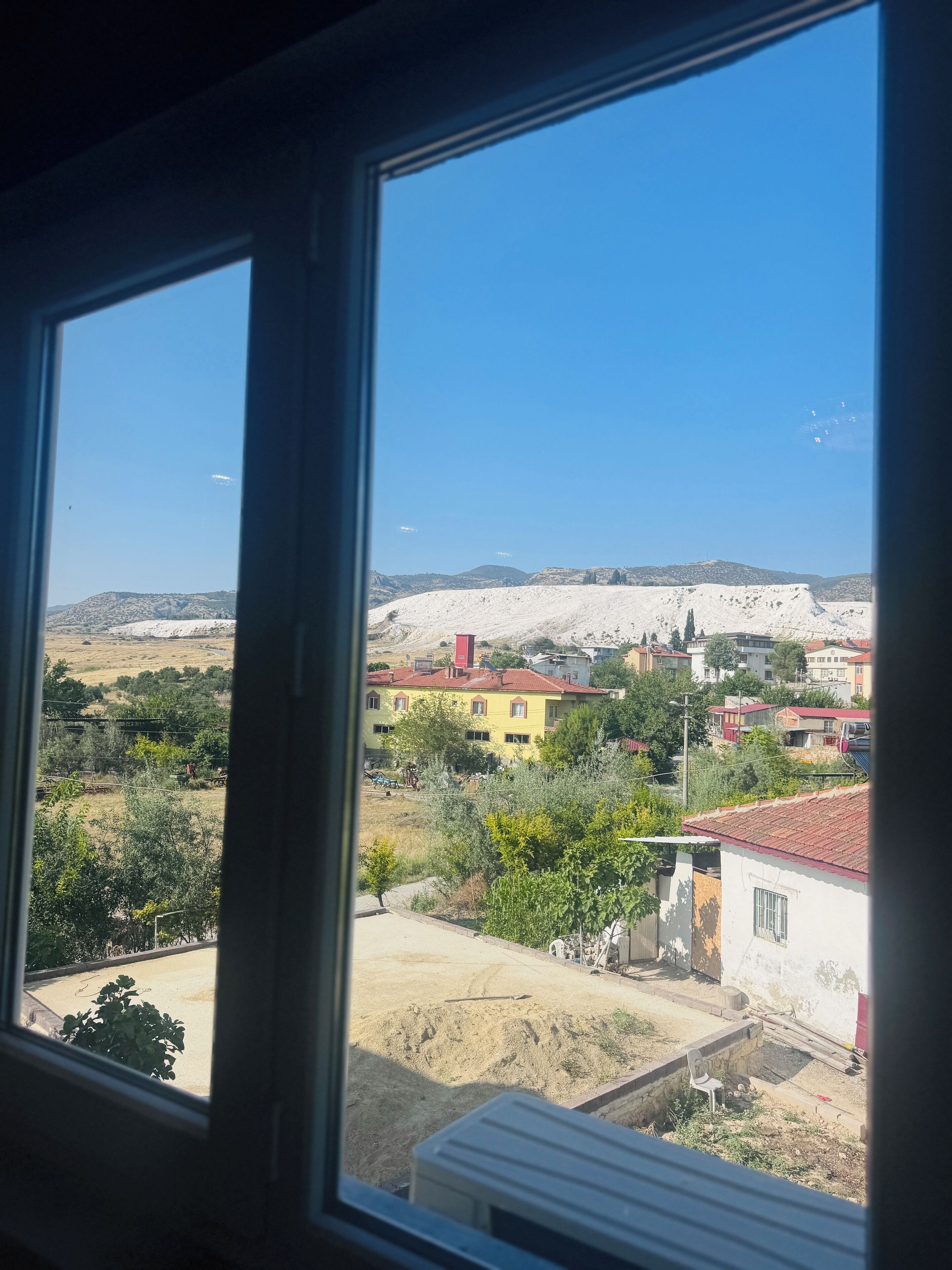 Vista pela janela de uma área residencial com casas, céu claro e uma cordilheira distante.