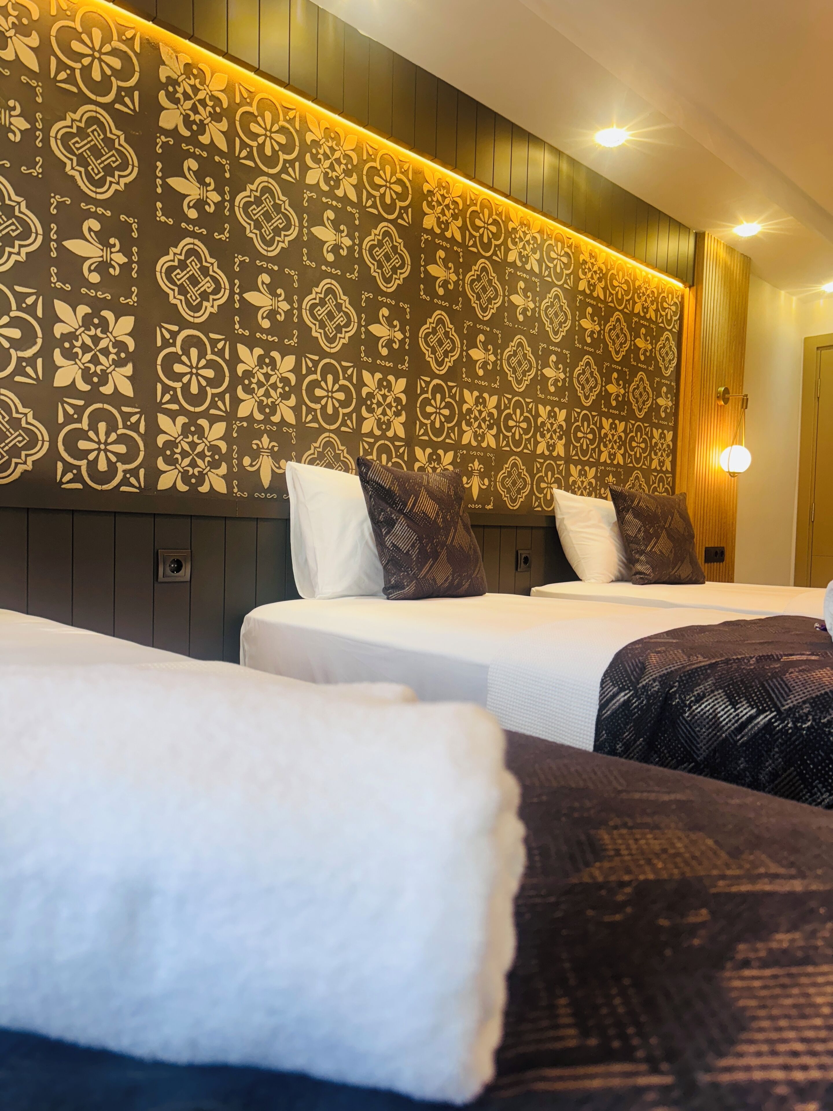 Habitación de hotel con dos camas, cada una con una colcha blanca y una manta estampada. La cabecera tiene un panel decorativo con diseños intrincados.