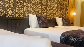 Habitación de hotel con dos camas, cada una con una colcha blanca y una manta estampada. La cabecera tiene un panel decorativo con diseños intrincados.